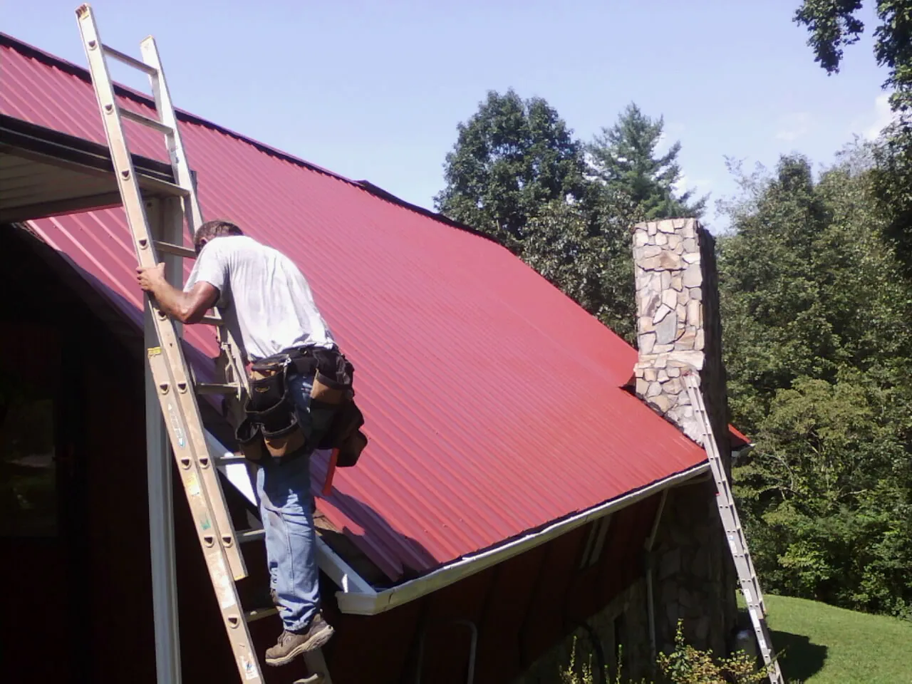 Roofer on a ladder beside a red metal roof and stone chimney — Soffit & Fascia Repair by Roofing Brothers in Paulina